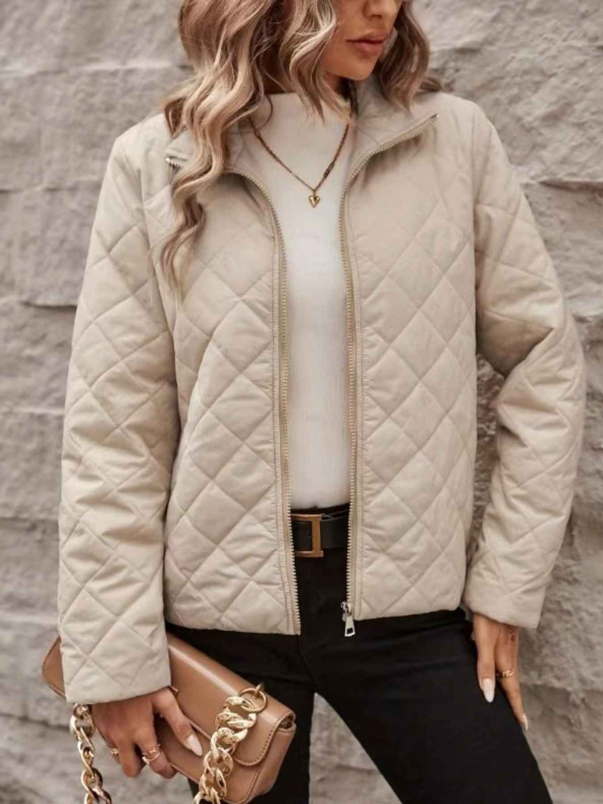 A woman wearing a quilted zip-up jacket in a light beige/cream color, styled with a white top, black pants, and a brown handbag, standing against a gray textured wall.