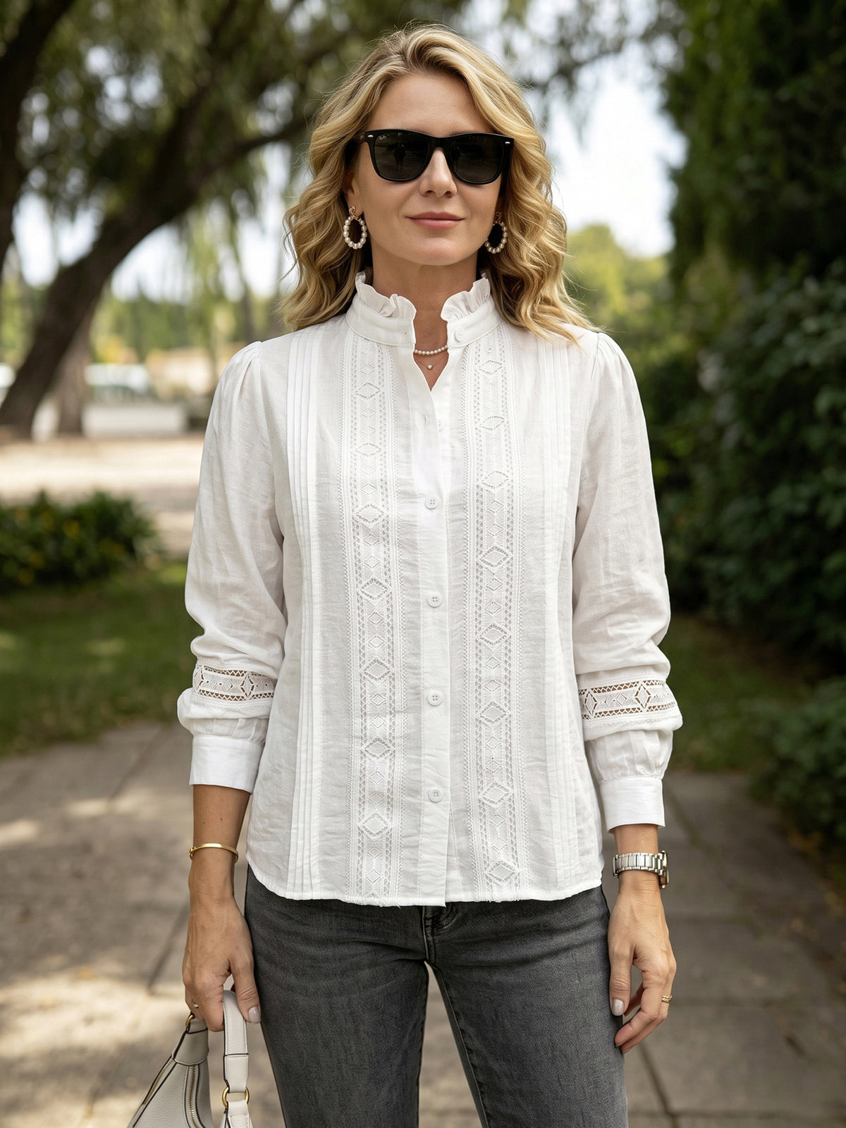 A woman wearing a white long-sleeve button-up blouse with vertical lace-like embroidery and lace cuffs, paired with dark gray jeans, standing outdoors on a paved path with trees in the background.