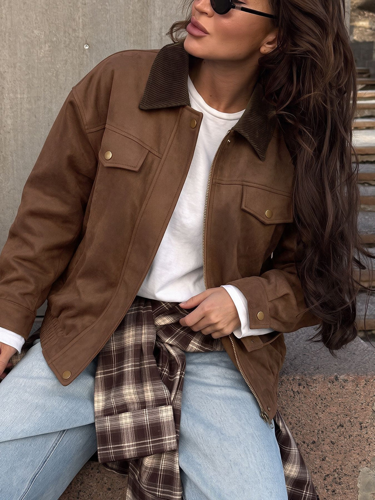 A woman wearing a brown suede jacket with a dark brown corduroy collar, styled over a white t-shirt, light blue jeans, and a brown plaid shirt tied around her waist, set against a concrete wall.