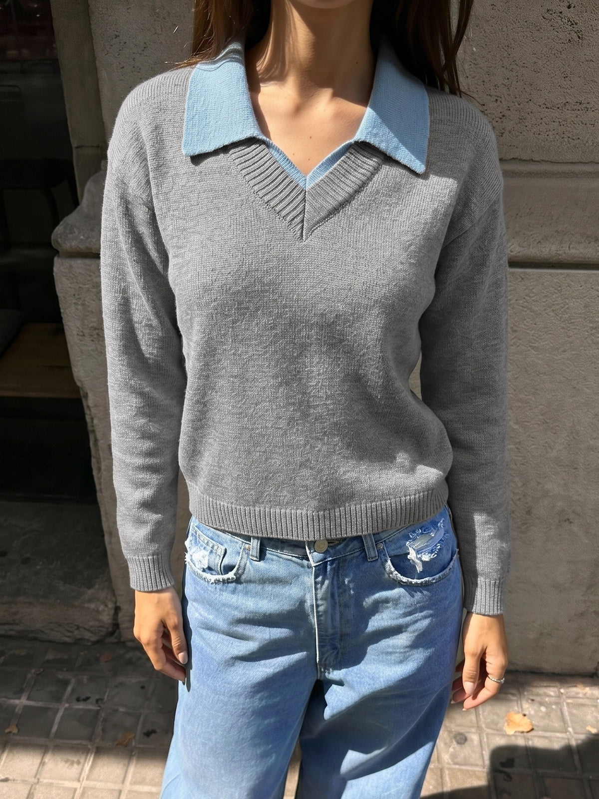 A woman wearing a light gray knit sweater with a contrasting light blue collar, paired with light blue jeans, standing outdoors against a stone wall.