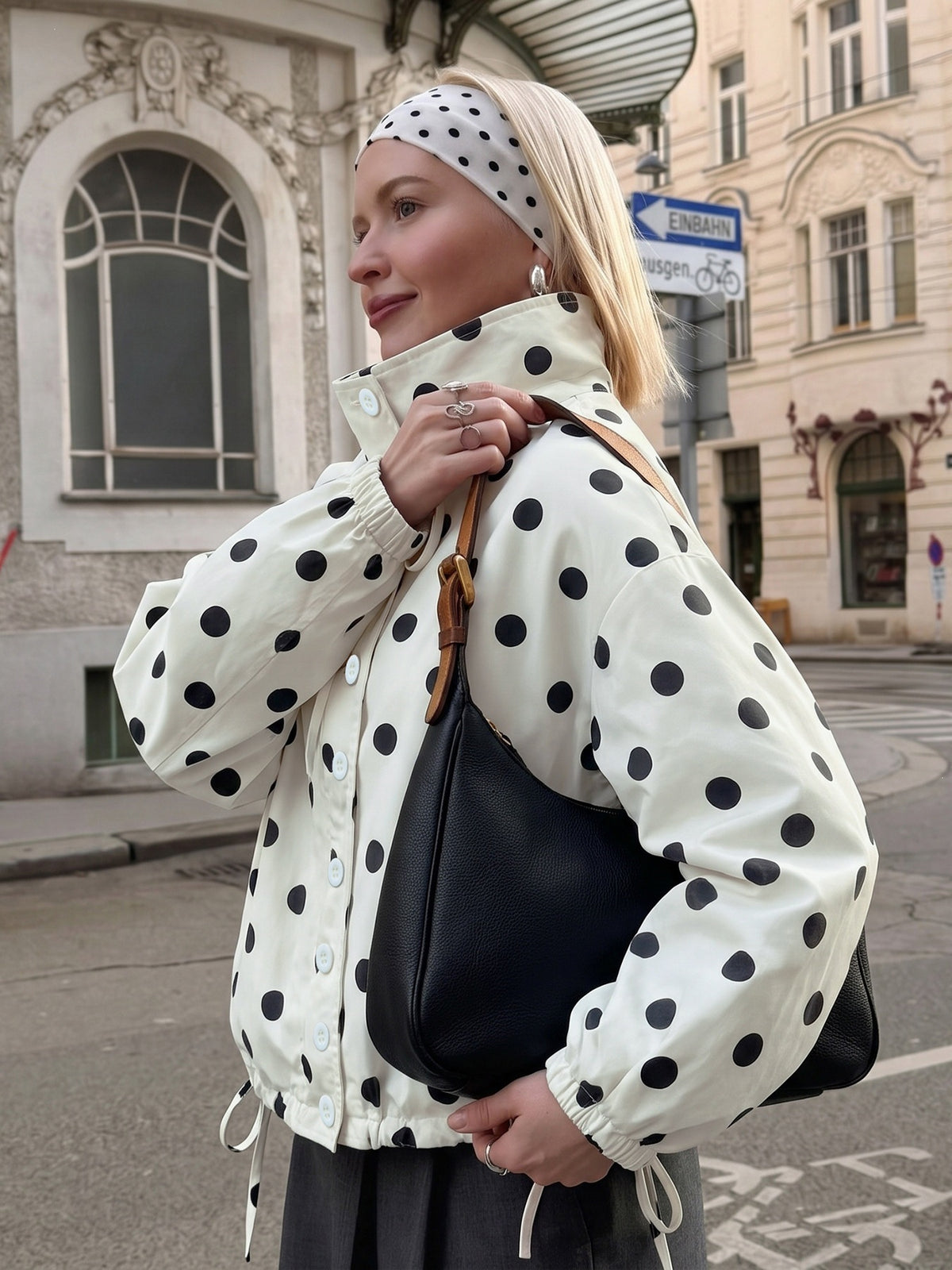 A woman wearing a cream-colored puffer jacket with black polka dots, a matching polka dot headband, and carrying a black shoulder bag, standing on a city street.