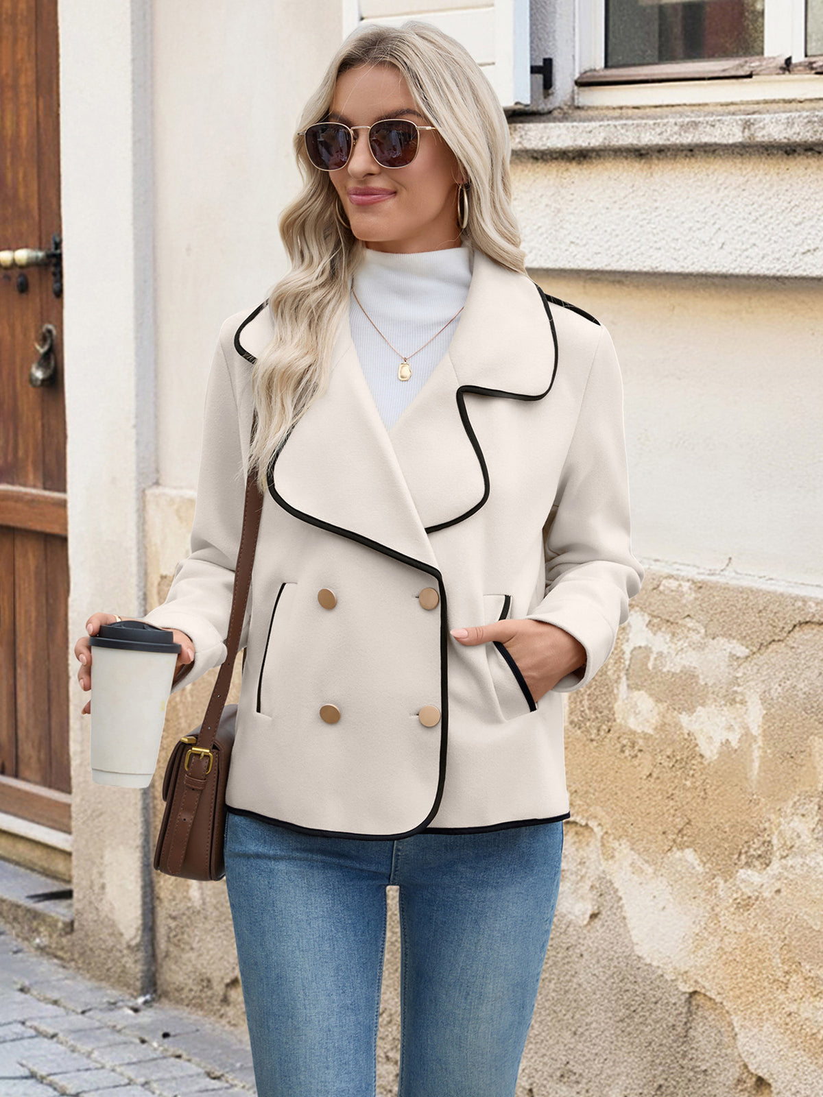 Woman wearing a stylish beige jacket with black trim, holding a coffee cup and a bag, standing against a building.
