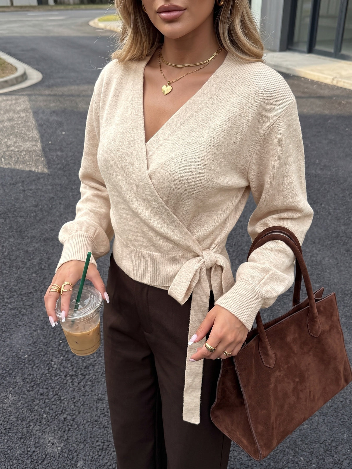 A woman wearing a beige knit wrap sweater with a tie waist, paired with dark brown pants, holding an iced coffee and carrying a brown suede tote bag.