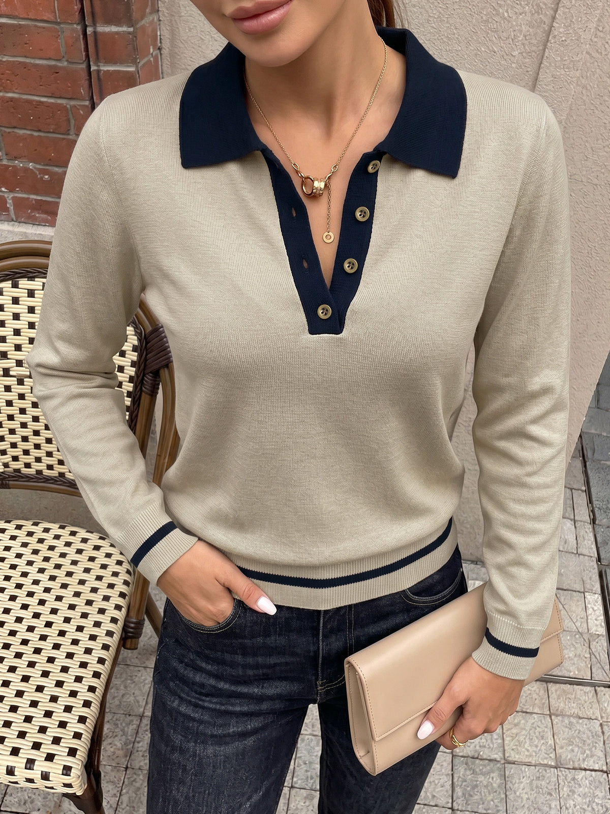 A woman wearing a beige long-sleeve polo sweater with a navy blue collar and trim, paired with dark blue jeans and holding a beige clutch purse.