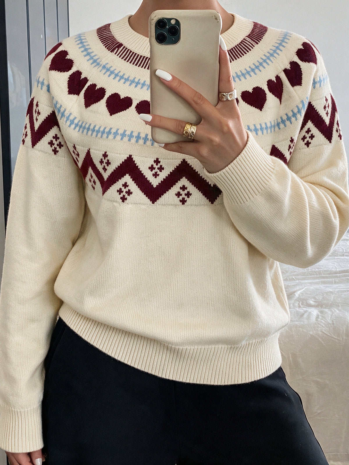 A person taking a mirror selfie wearing a cream-colored knit sweater with a Fair Isle pattern of burgundy hearts and geometric motifs, paired with black pants.