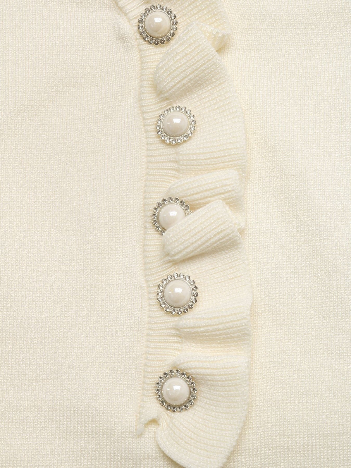 Close-up of a cream-colored ribbed knit top featuring a vertical ruffle trim adorned with five decorative buttons that have white faux pearls and silver rhinestone borders.