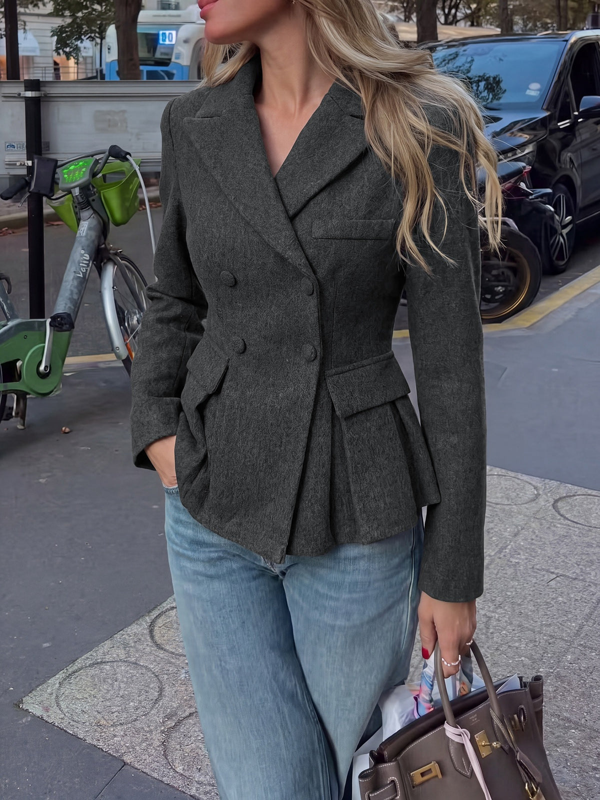 A woman wearing a dark olive green double-breasted blazer with a peplum hem, paired with light blue jeans and holding a brown handbag on a city street.