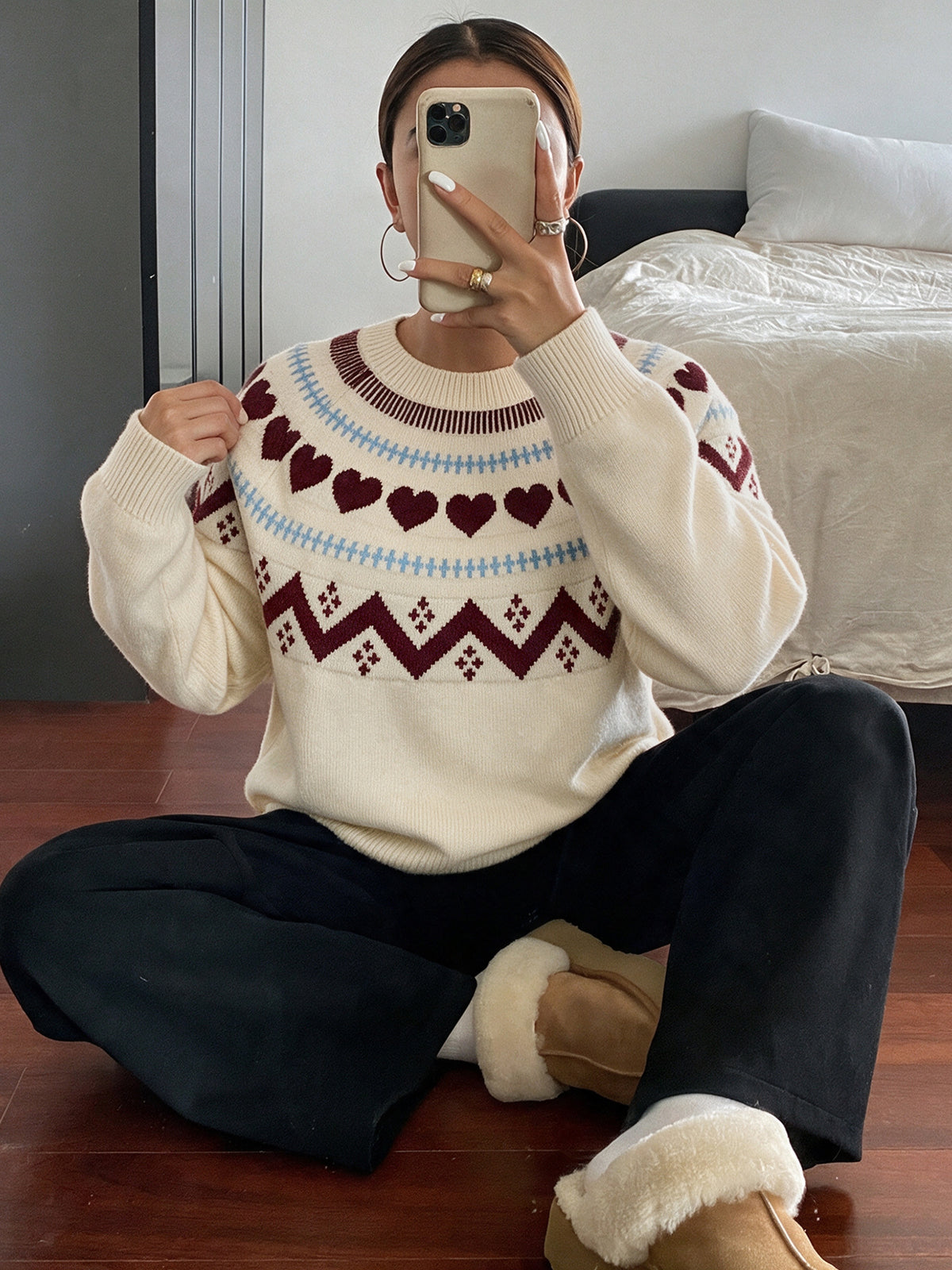 A woman takes a mirror selfie wearing a cream-colored knit sweater with a Fair Isle pattern of red hearts and burgundy geometric motifs, paired with black pants and tan slippers with white faux fur trim.