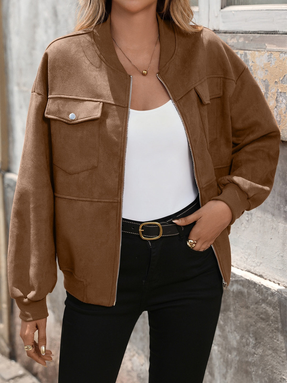 Person wearing a brown jacket over a white top and black pants, standing against an outdoor background.