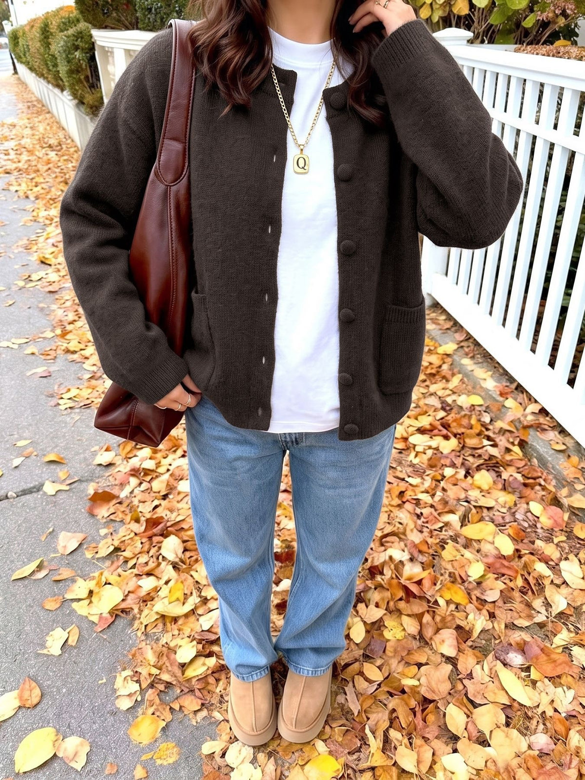 A person standing outdoors on a path covered with fallen autumn leaves, wearing a dark brown button-front cardigan over a white T-shirt, light blue jeans, tan boots, and carrying a brown shoulder bag.