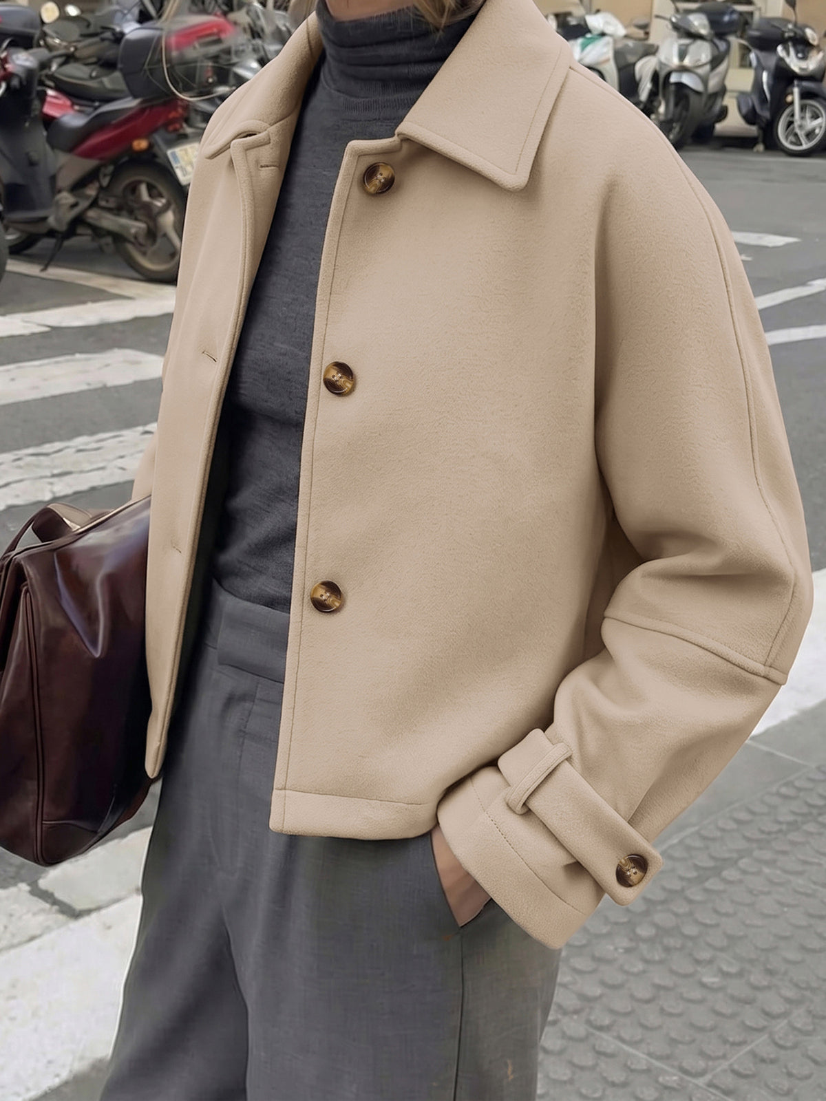 A person wearing a beige oversized coat over a dark gray turtleneck and gray trousers, standing on a city street with parked scooters in the background.