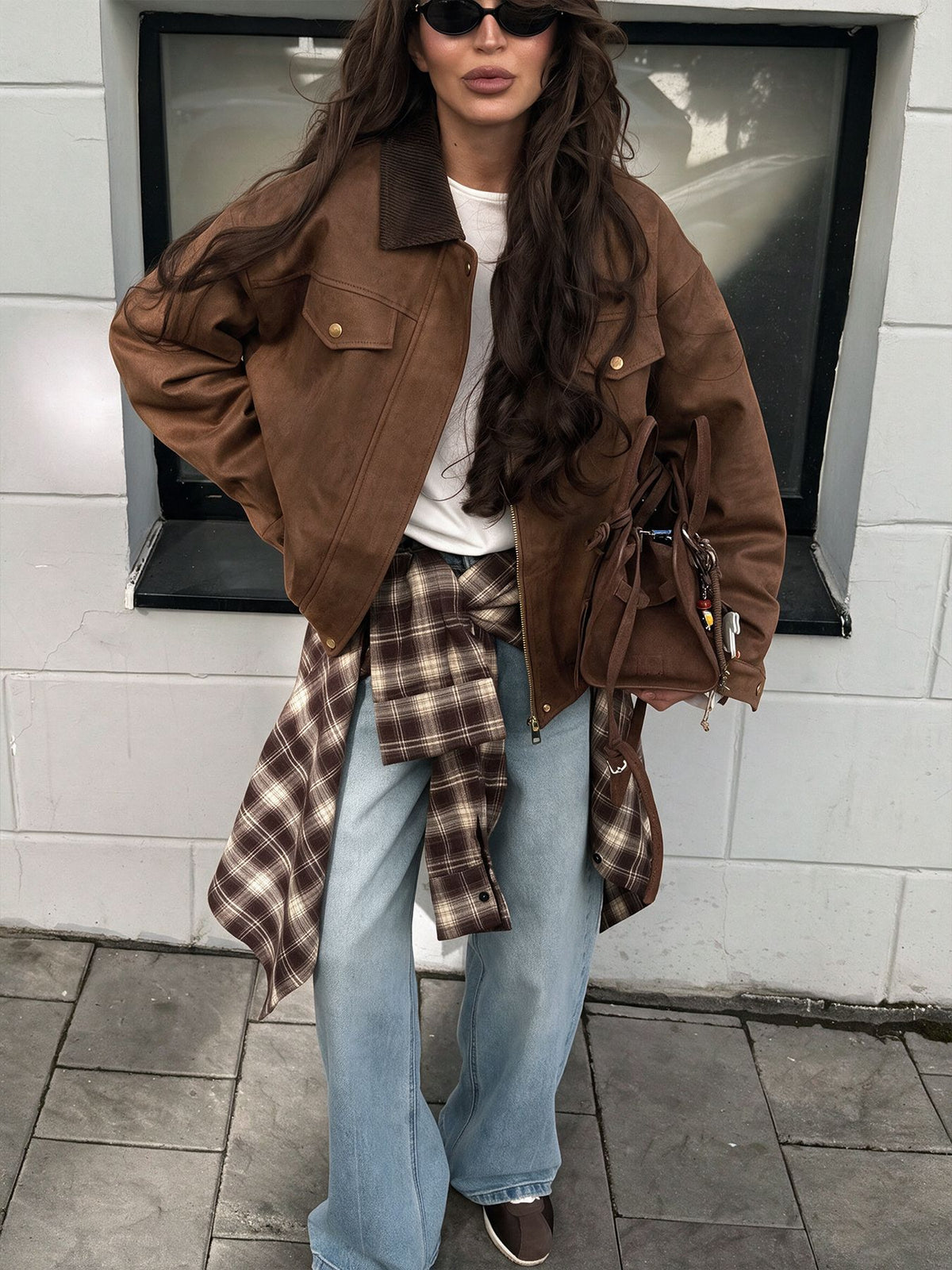 A woman wearing a brown suede jacket over a white t-shirt, light blue jeans, and a brown plaid shirt tied around her waist, carrying a matching brown shoulder bag.