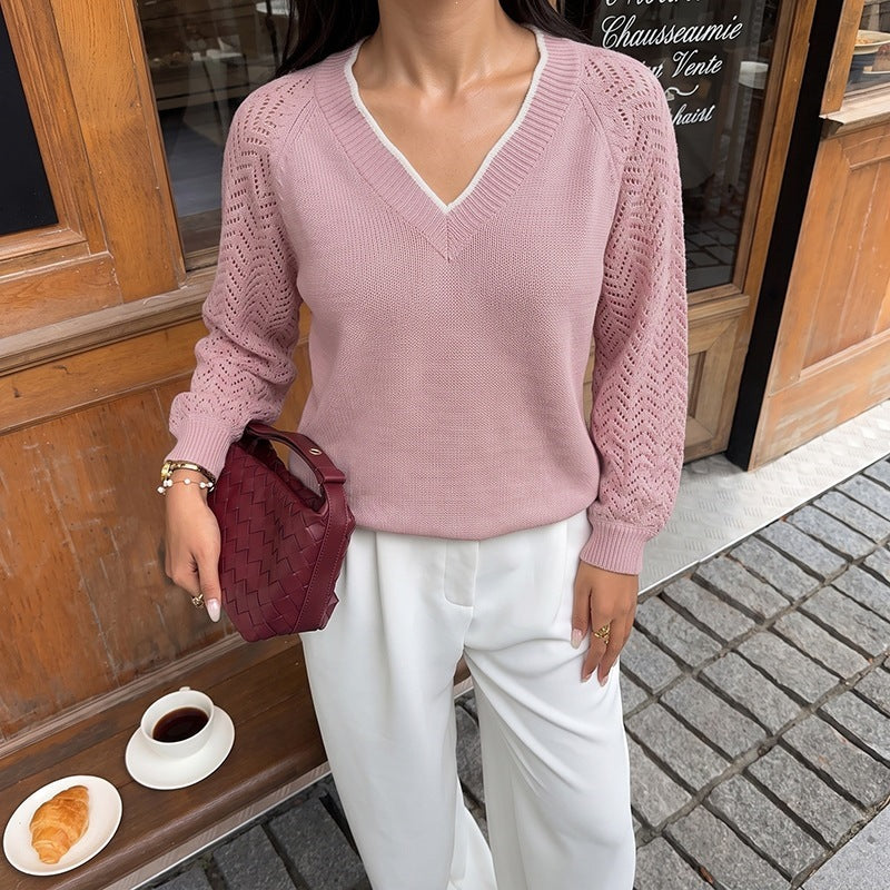 A woman wearing a pink V-neck knit sweater with textured sleeves, paired with white trousers and holding a burgundy woven handbag, standing on a cobblestone street in front of a wooden storefront.