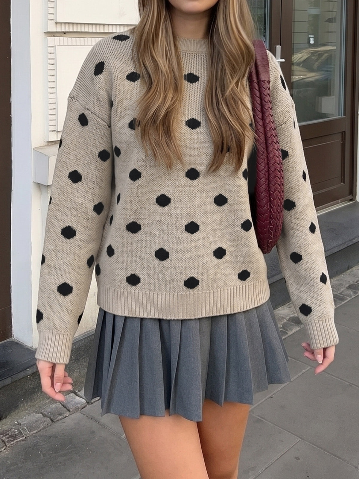 A woman wearing a beige knit sweater with black polka dots and a gray pleated mini skirt, carrying a burgundy woven shoulder bag, standing outdoors on a sidewalk.