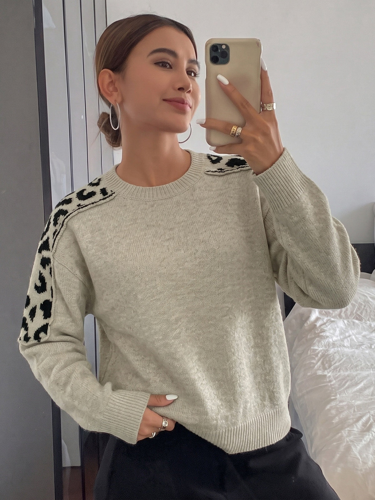 A woman takes a mirror selfie wearing a light beige crewneck sweater with black leopard print details on the shoulders and sleeves, paired with black pants.