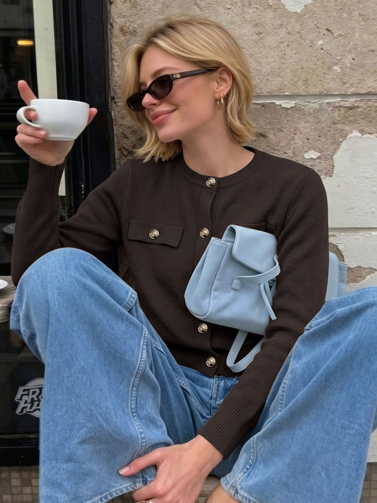 A woman wearing a dark brown ribbed cardigan with gold buttons, light blue wide-leg jeans, and a light blue crossbody bag, sitting against a stone wall and holding a white coffee cup.