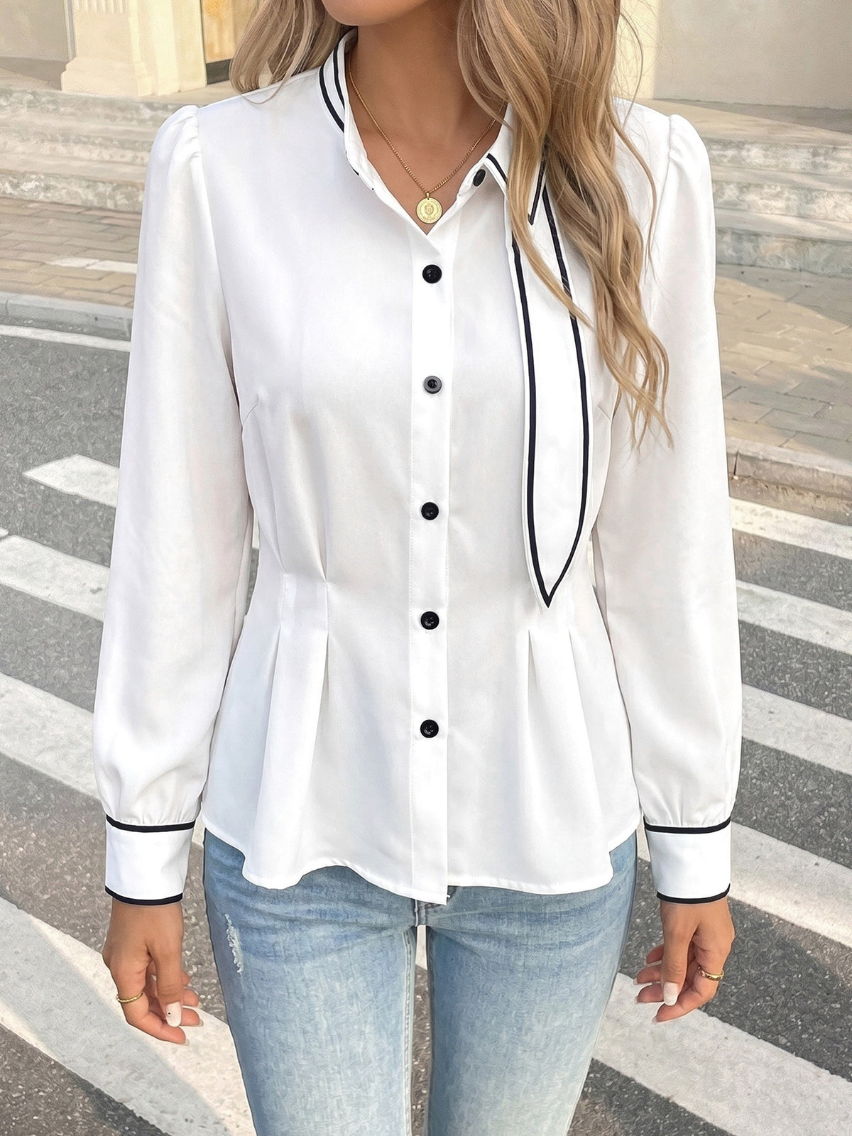 A woman wearing a white long-sleeve button-up blouse with black piping trim and a tie detail at the collar, paired with light blue jeans, standing outdoors on a crosswalk.