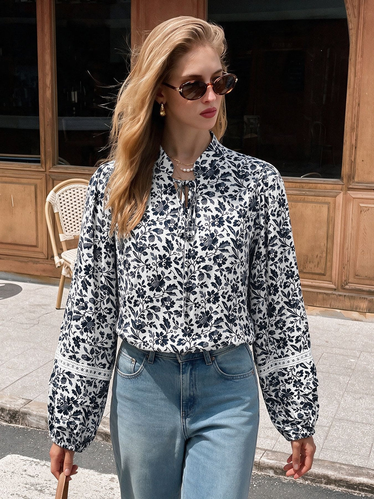 A woman wearing a long-sleeve white blouse with a black floral print and lace-up tie collar, paired with light blue jeans, walking outdoors in front of a wooden building.