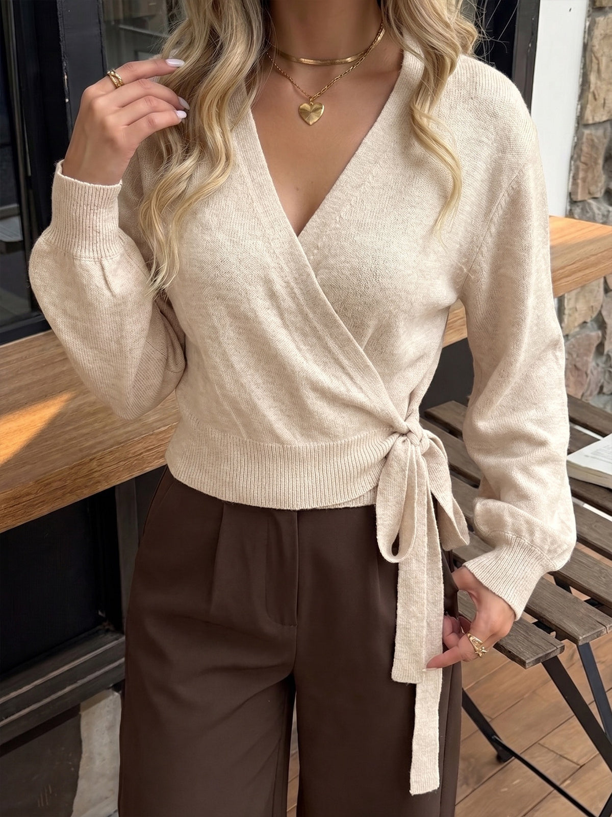 A woman wearing a beige knit wrap sweater with a tie waist and brown wide-leg pants, standing in a real indoor-outdoor setting with wooden furniture and a stone wall visible.