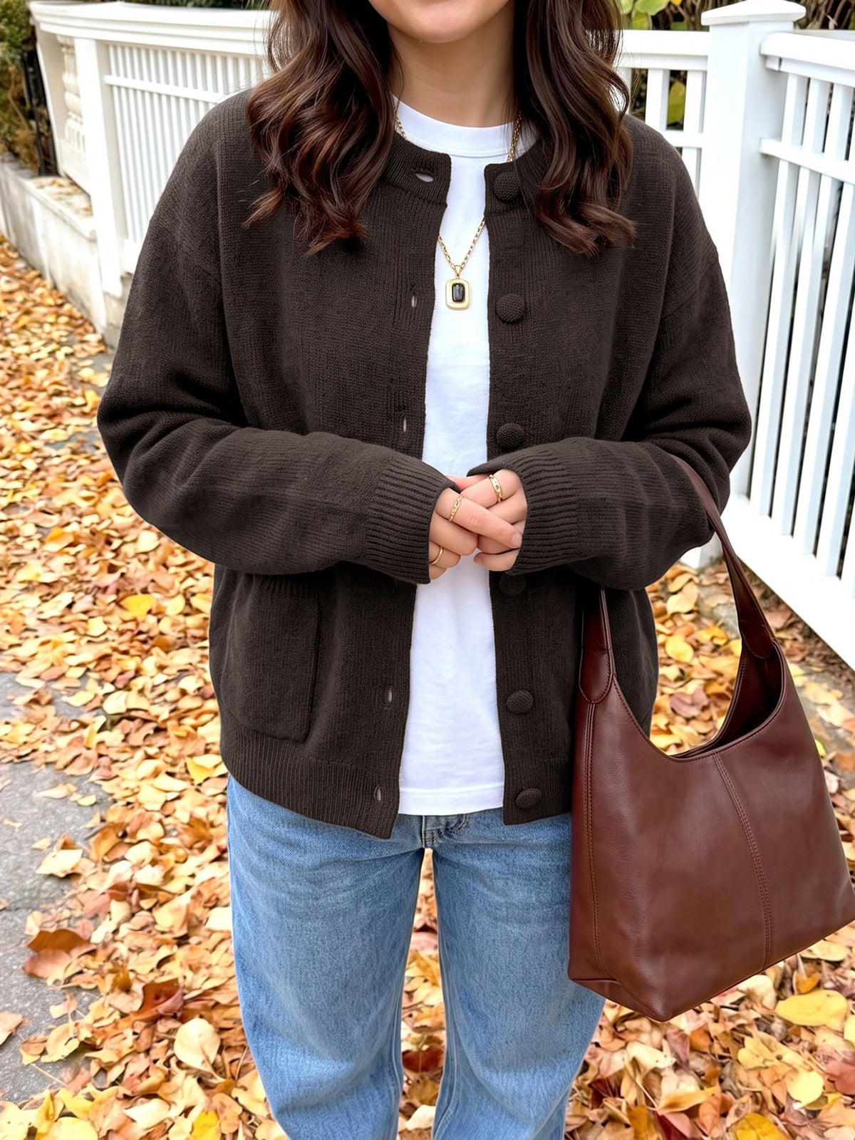 A person wearing a dark brown button-front knit cardigan over a white T-shirt and blue jeans, standing outdoors on a path covered with fallen leaves next to a white fence. A brown leather shoulder bag is also visible.