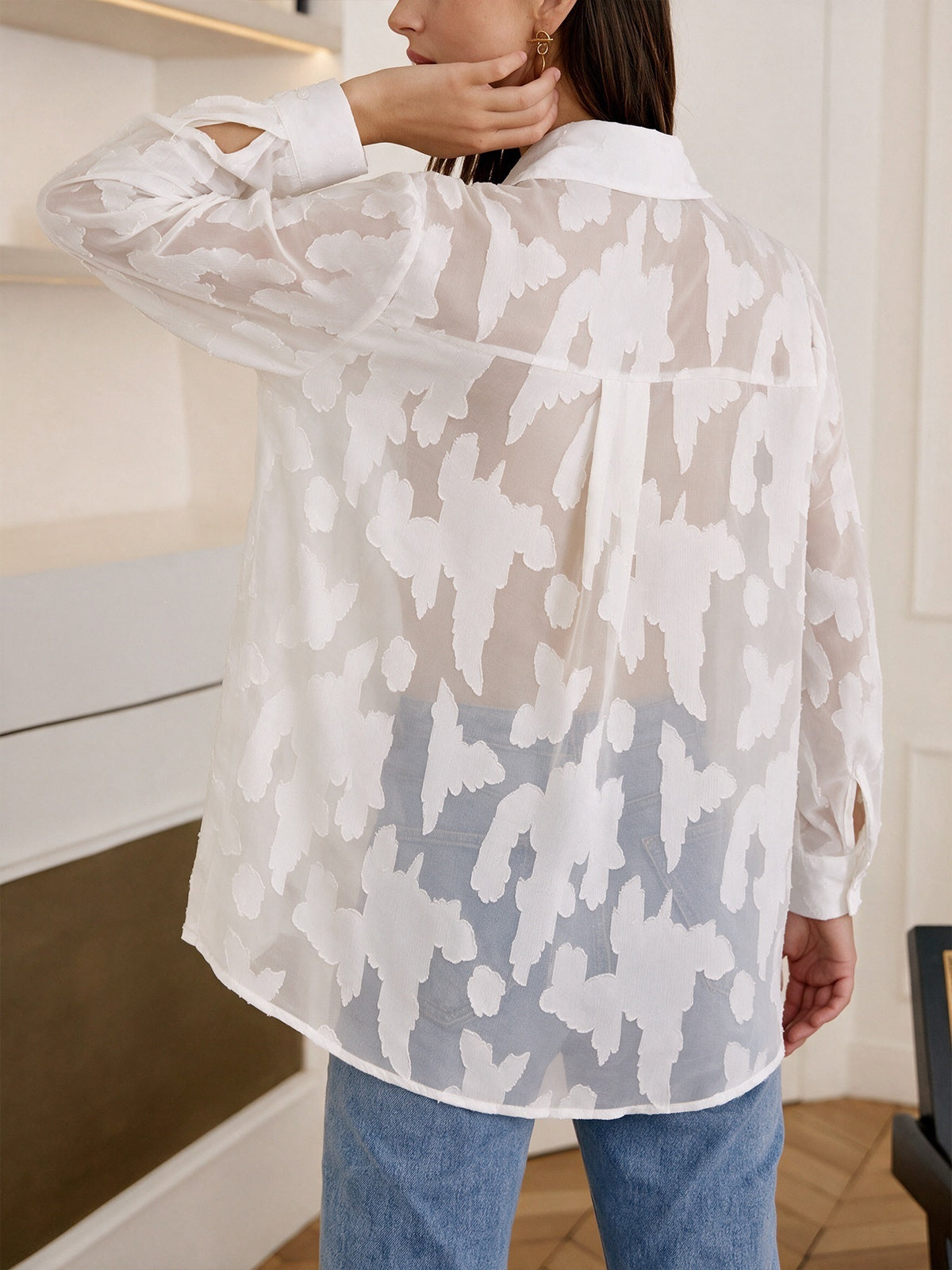 A woman wearing a sheer white long-sleeve blouse with an abstract white-on-white floral pattern, paired with blue jeans, shown from the back in an indoor setting.