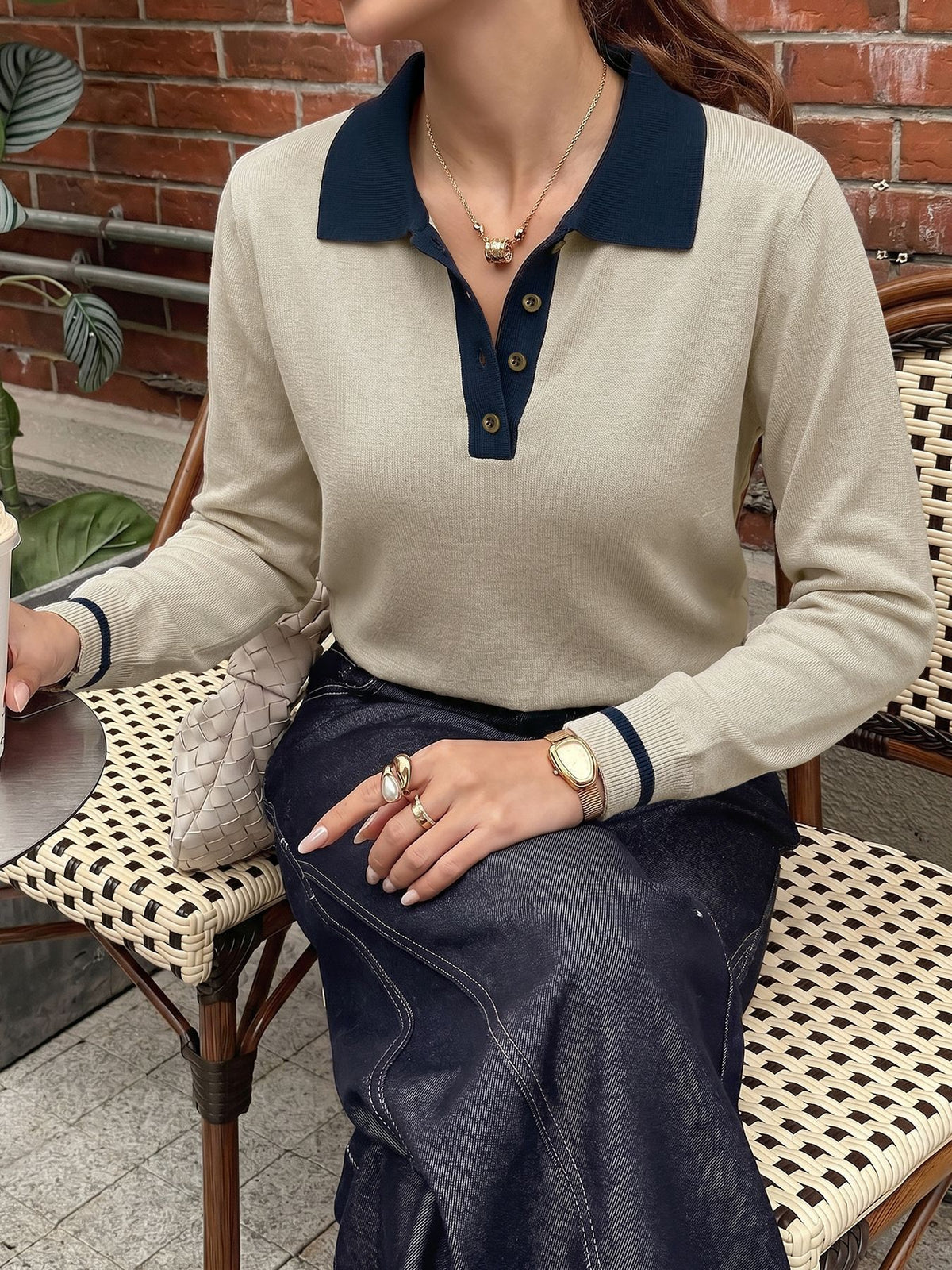 A woman seated on a woven chair wearing a beige long-sleeve polo sweater with a dark navy collar and button placket, paired with a dark denim skirt.