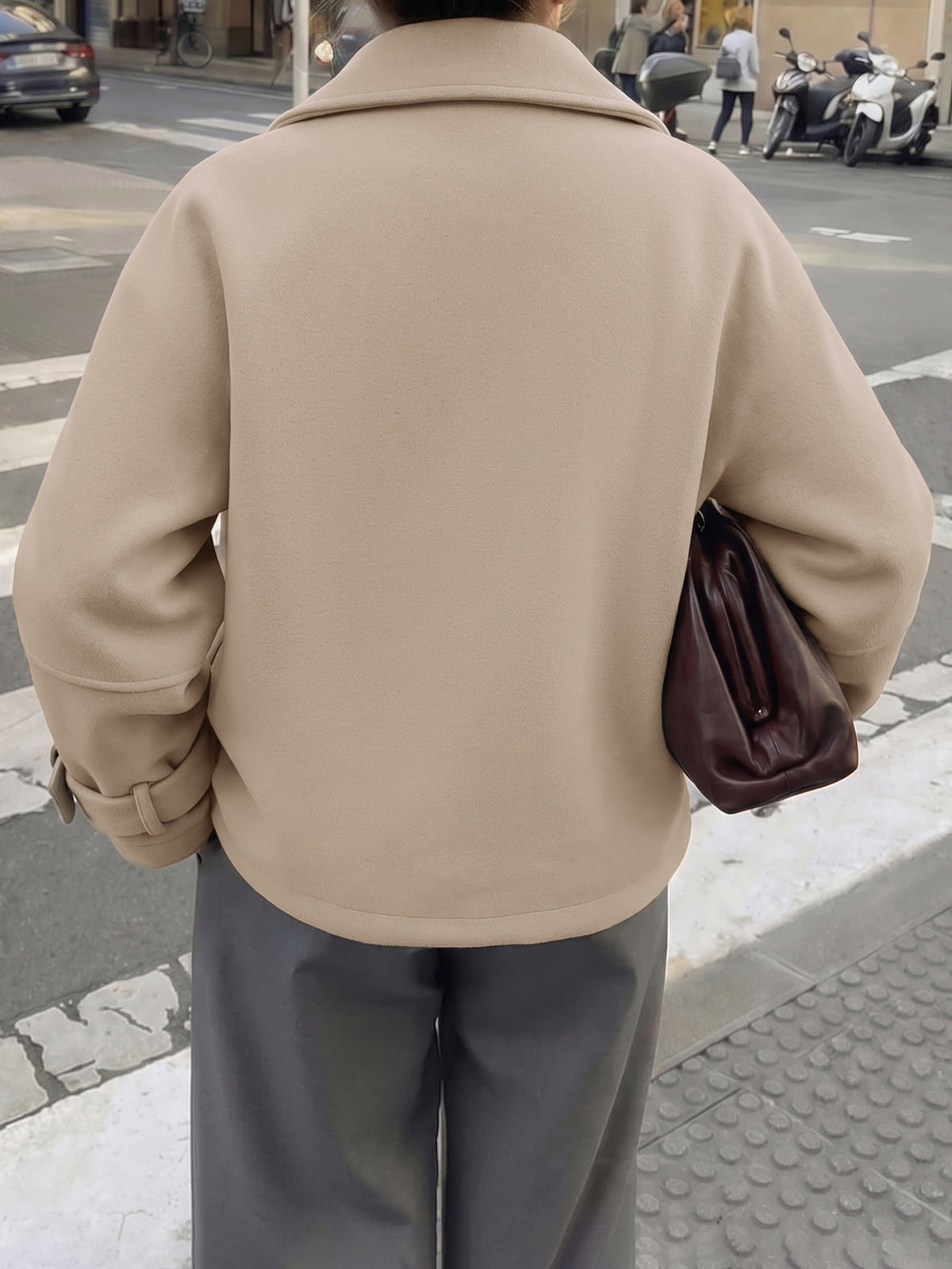 A person viewed from behind wearing a beige oversized coat and gray wide-leg pants, carrying a dark brown shoulder bag on a city street.
