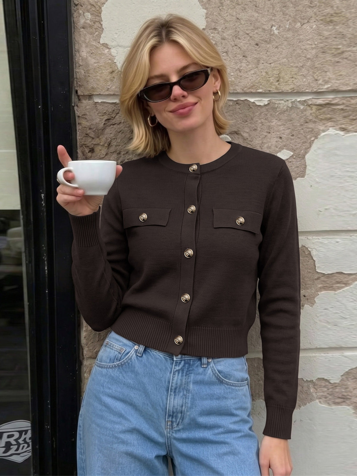 A woman wearing a dark brown ribbed cardigan with gold-tone buttons and flap chest pockets, paired with light blue jeans and sunglasses, holding a white mug against a stone wall background.