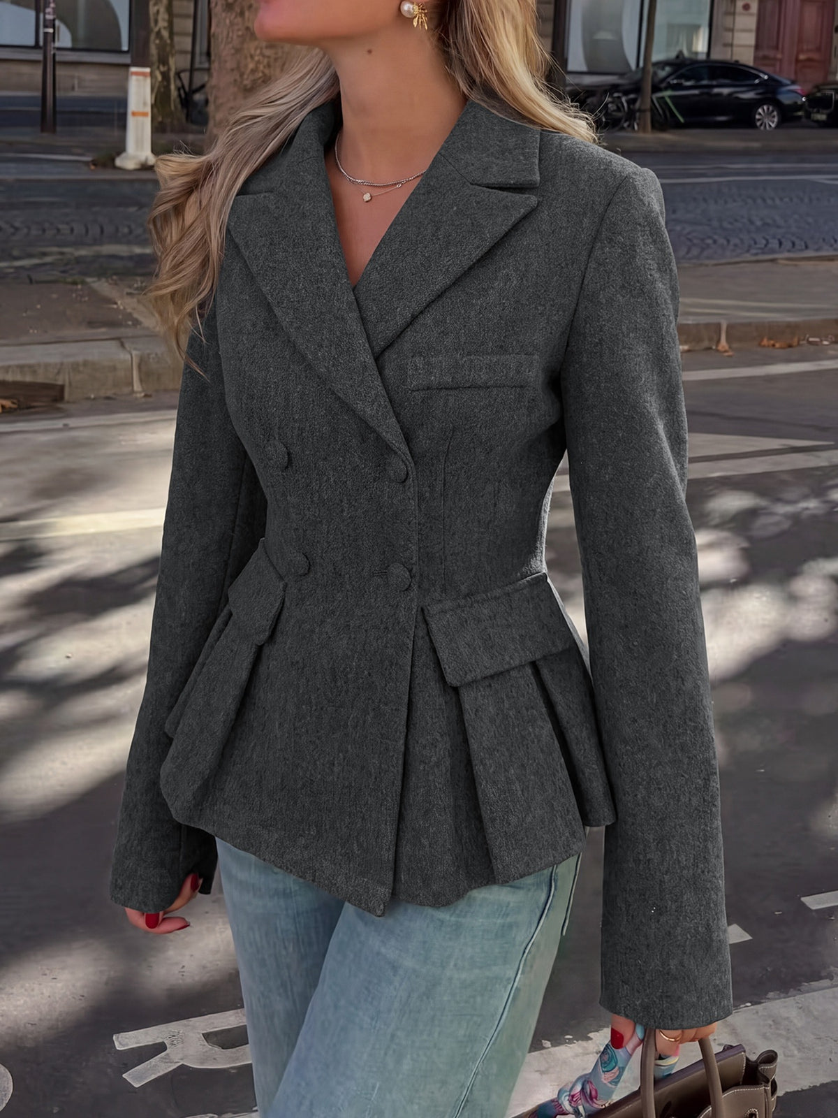 A woman wearing a dark gray double-breasted blazer with a peplum waist and flap pockets, paired with light blue jeans, standing on a city street.