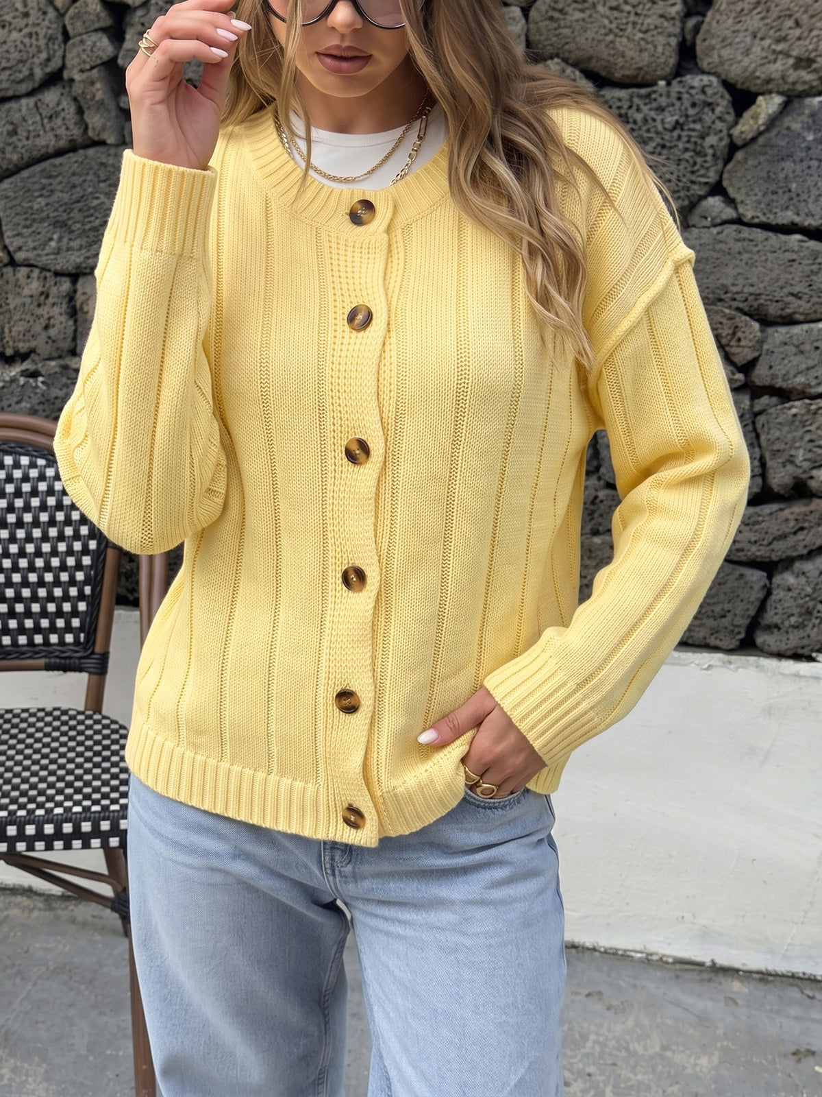 A woman wearing a light yellow cable-knit cardigan sweater with brown buttons, styled with light blue jeans, standing in front of a dark stone wall.