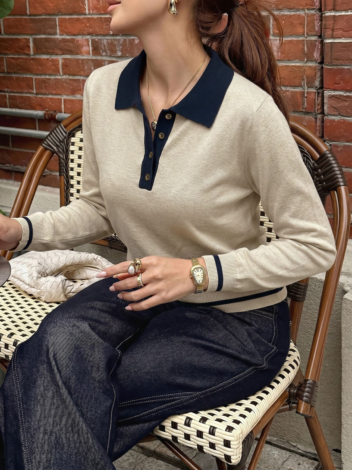 A woman wearing a beige long-sleeve polo sweater with a dark navy collar and button placket, paired with dark blue jeans, sitting on a woven rattan chair against a red brick wall.
