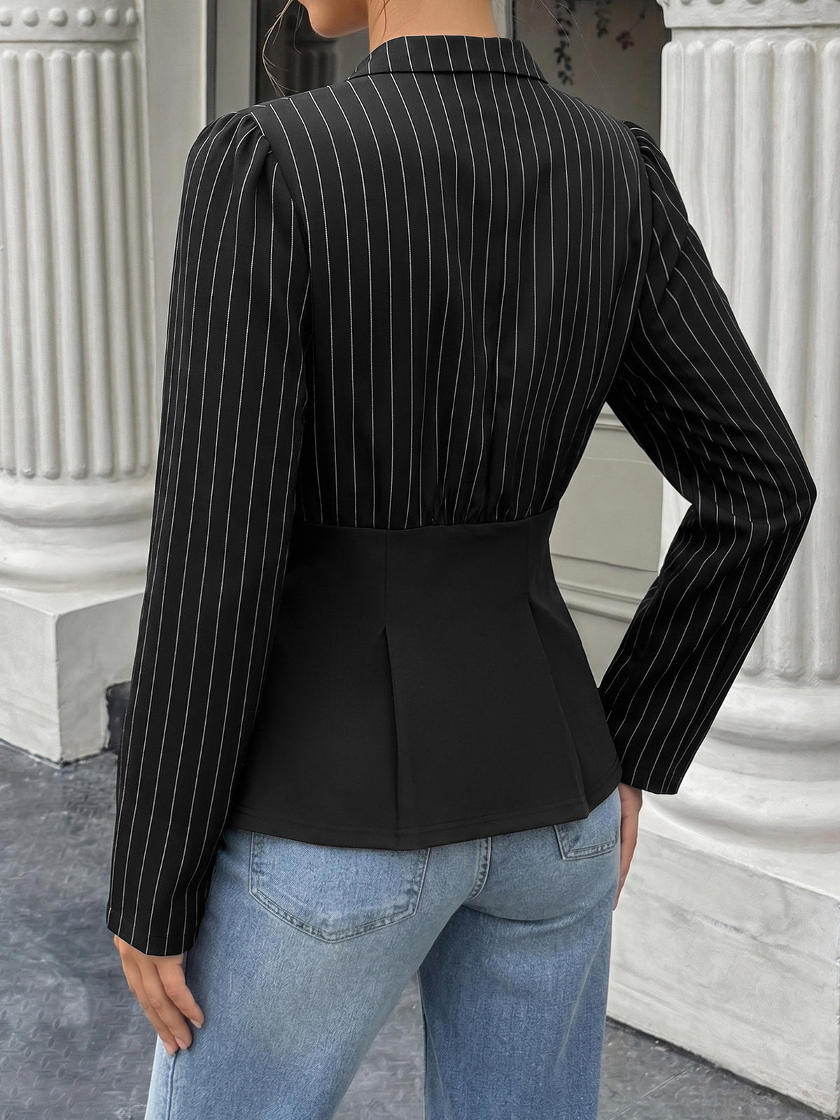 Person wearing a black pinstripe blazer and blue jeans standing in front of classical architecture.