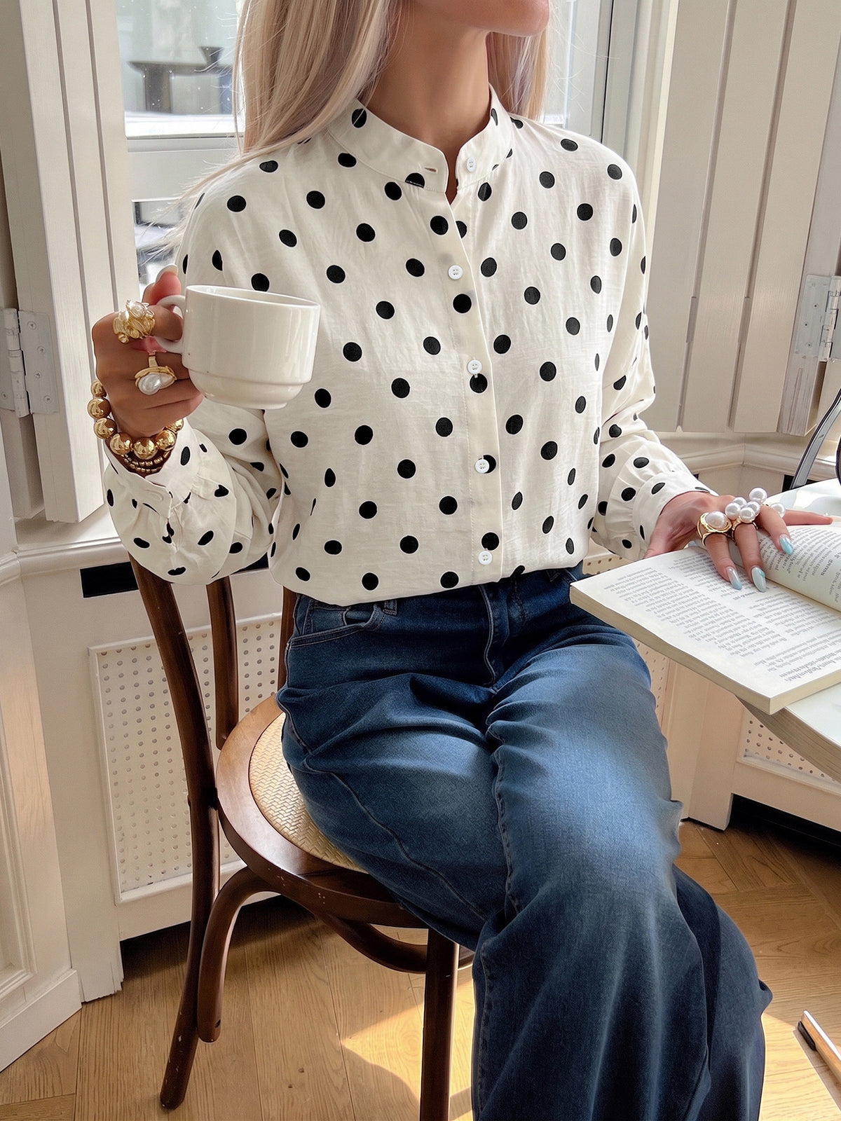 A woman seated on a wooden chair indoors wearing a white long-sleeve blouse with black polka dots and blue jeans, holding a white mug and an open book.