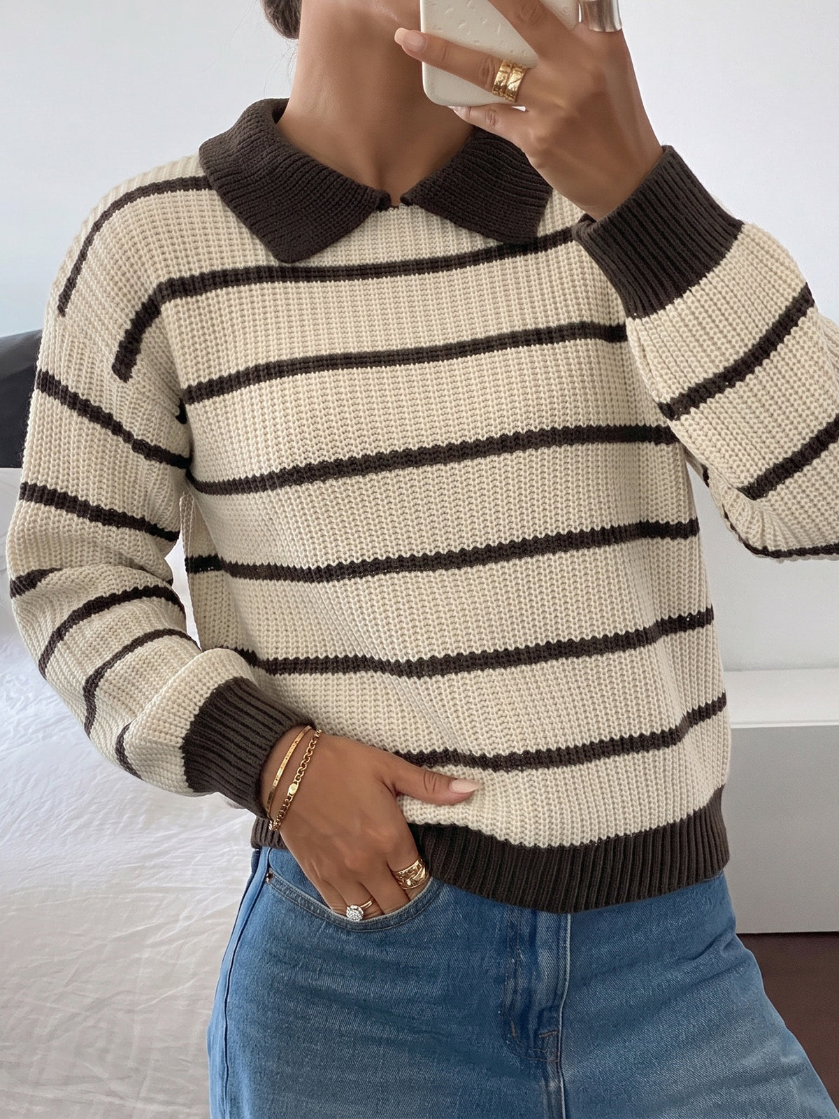 A person wearing a cream and dark brown horizontally striped knit sweater with a dark brown collar and ribbed cuffs and hem, paired with blue jeans.