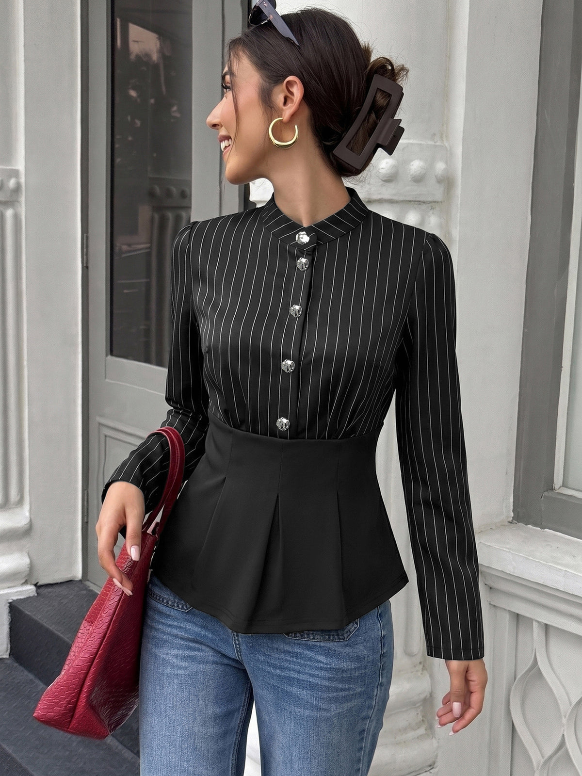 A woman wearing a black long-sleeve blouse with thin white vertical pinstripes and a buttoned stand collar, paired with blue jeans and holding a red handbag, standing in front of a white building entrance.