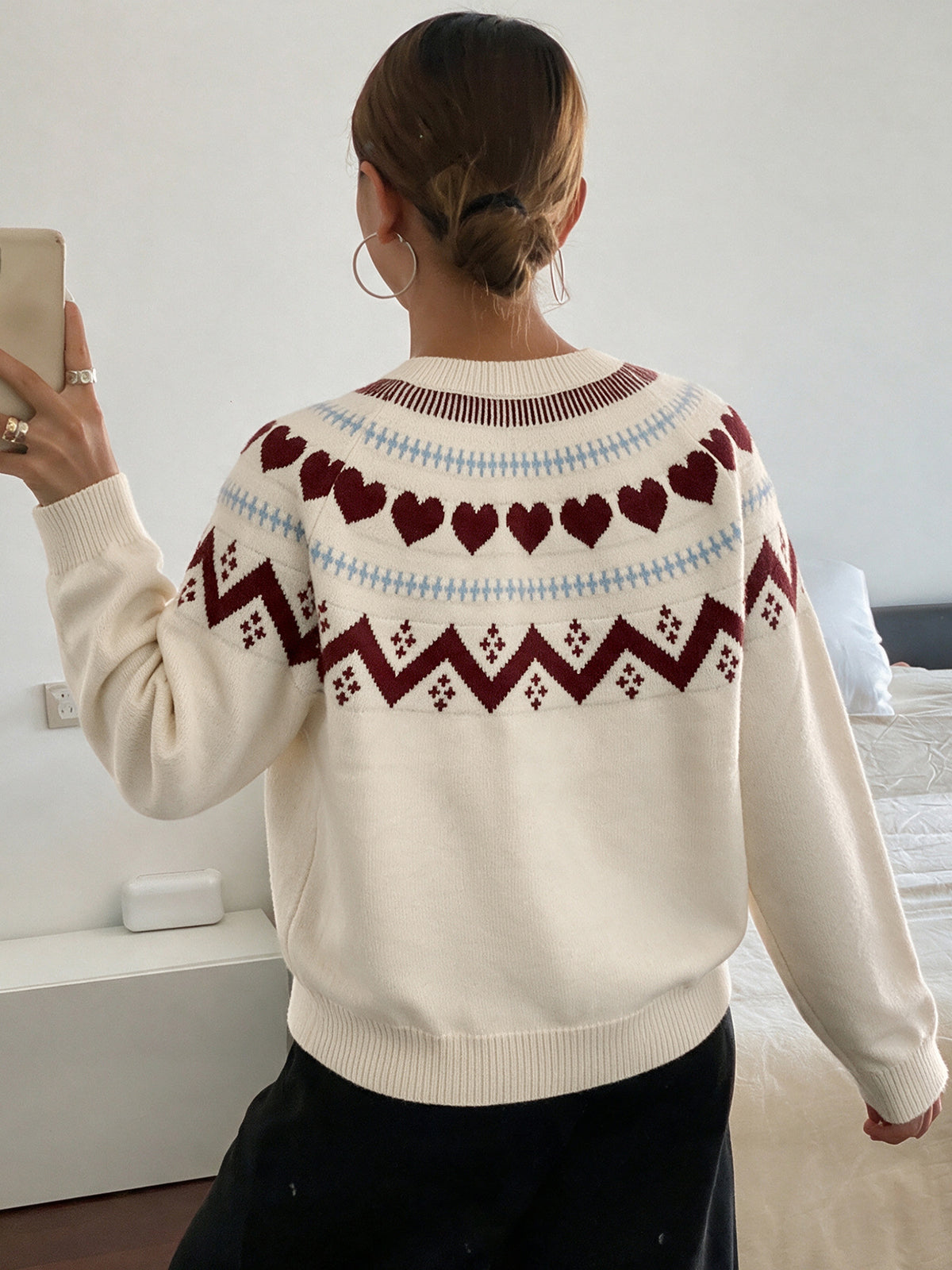A woman viewed from behind wearing a cream-colored knit sweater with a Fair Isle pattern of red hearts and geometric designs, paired with black pants in a bedroom setting.