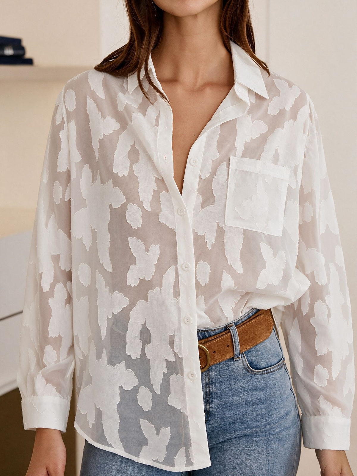 A woman wearing a sheer white button-up blouse with an abstract white-on-white leopard print, tucked into blue jeans with a brown belt.