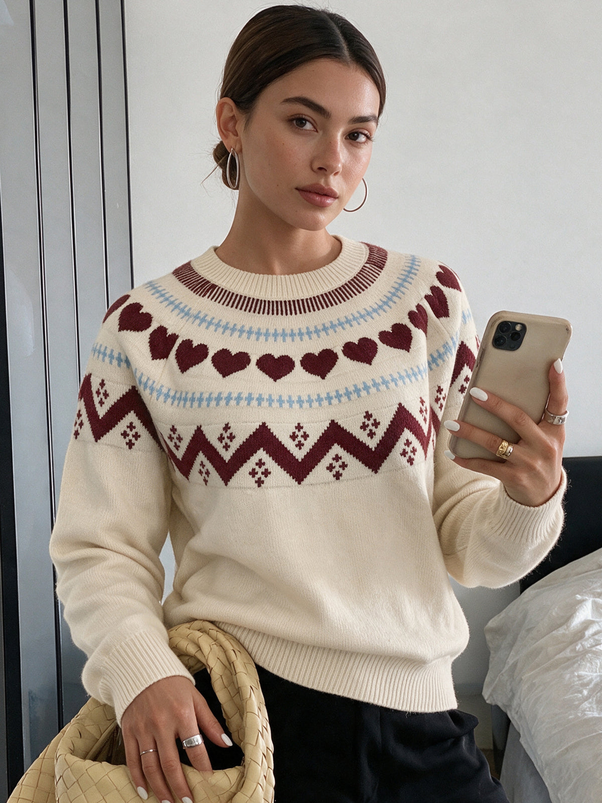 A woman takes a mirror selfie wearing a cream-colored knit sweater with a Fair Isle pattern of dark red hearts and geometric motifs, accented with light blue stripes.
