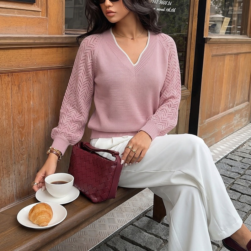 A woman sits on a wooden bench wearing a light pink V-neck knit sweater with textured sleeves, paired with white pants and a burgundy woven handbag. A cup of coffee and a croissant are on the table in front of her, set against a rustic wooden storefront background.
