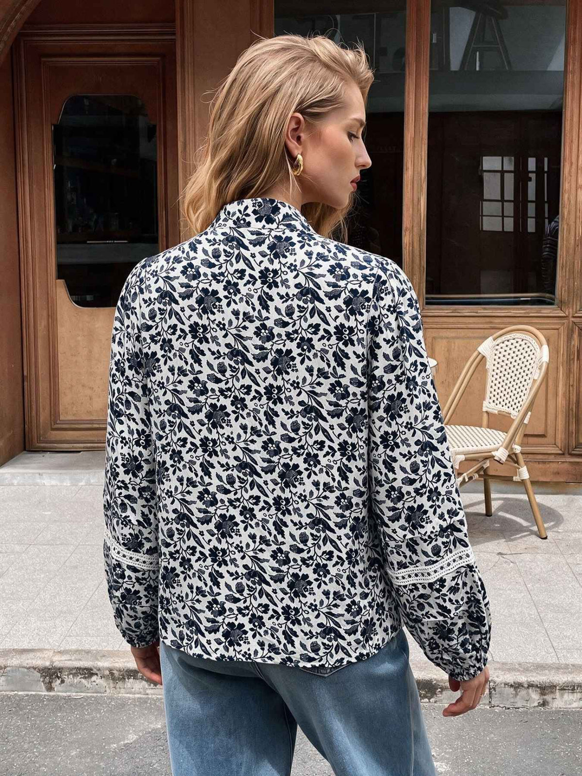 A woman viewed from behind wearing a long-sleeve floral blouse with a high collar and blue jeans, standing outdoors in front of a wooden building.