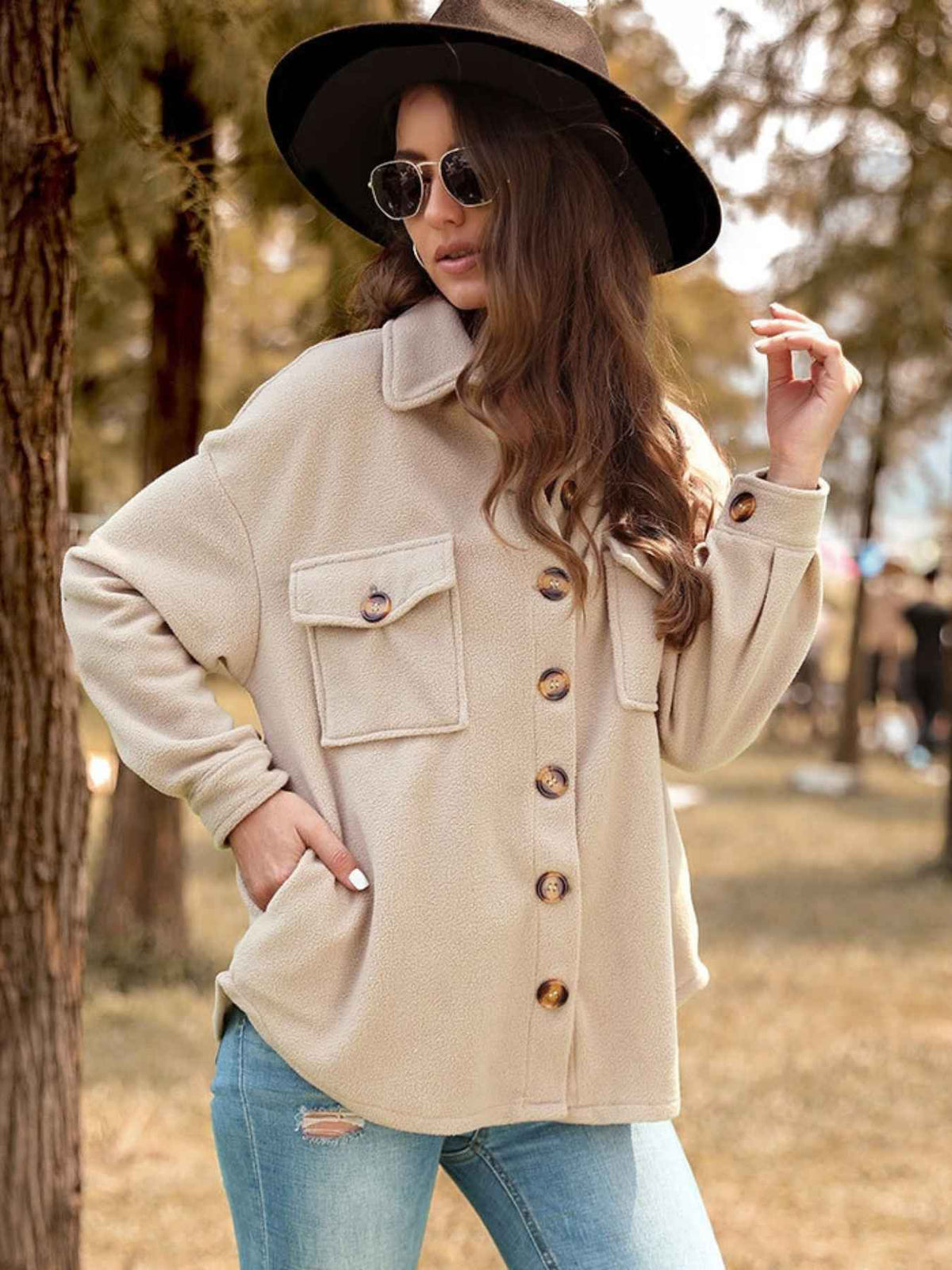 A woman wearing a beige oversized button-front jacket with flap chest pockets, blue jeans, a wide-brim black hat, and sunglasses, standing outdoors in a park-like setting with trees and dry grass.