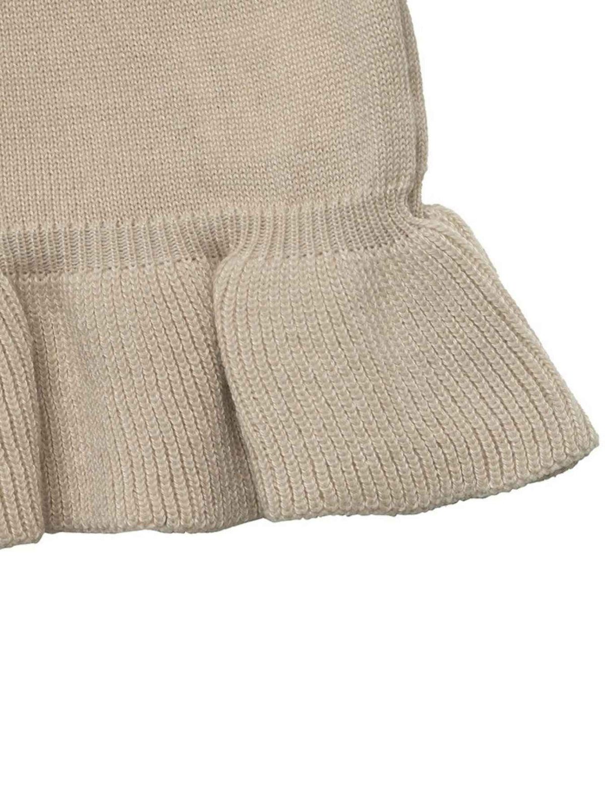 Close-up of a ribbed knit garment in a light warm beige or oatmeal color, showing the texture and ruffled hem detail against a plain white background.