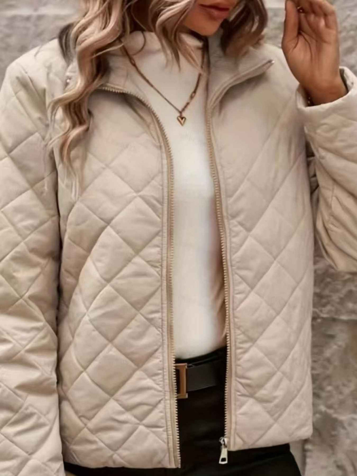 A close-up of a woman wearing a quilted zip-up jacket in a light cream or off-white color, layered over a white top and black pants.