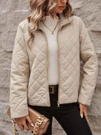 A woman wearing a quilted zip-up jacket in a light beige/cream color, styled with a white top, black pants, and a brown handbag, standing against a gray textured wall.