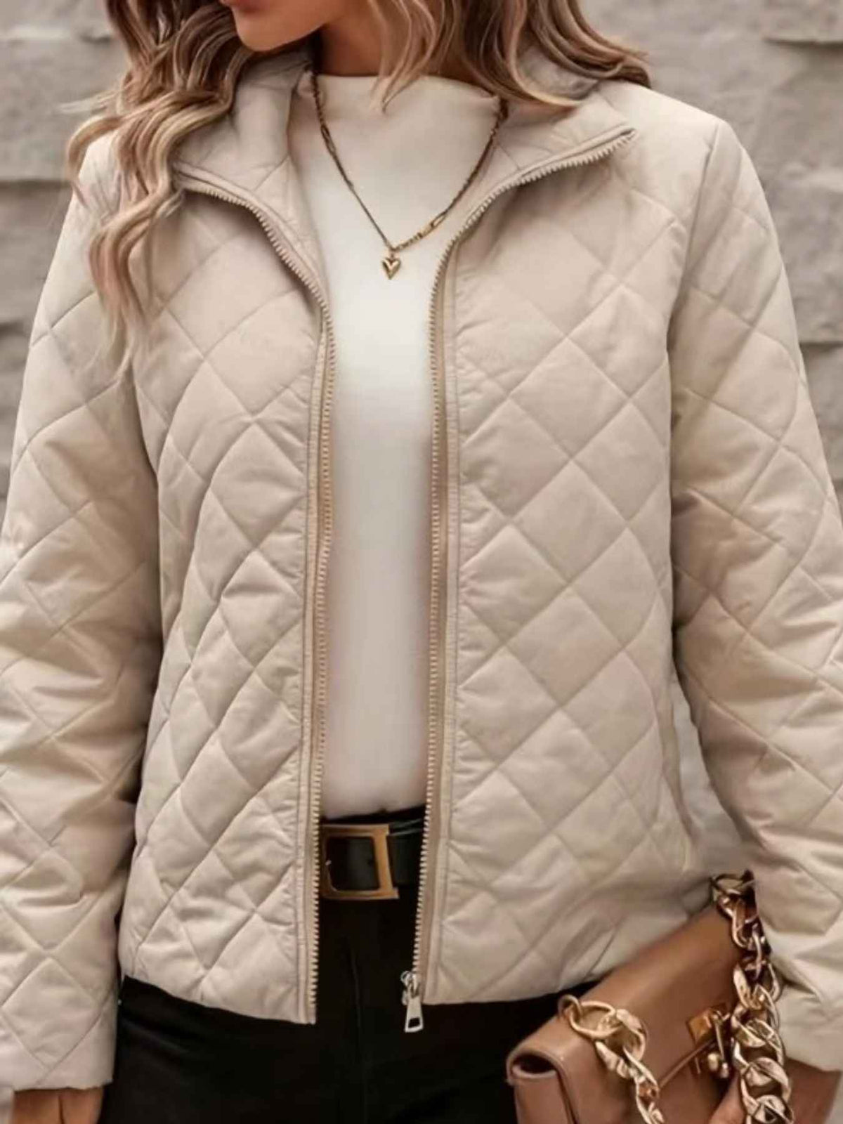A woman wearing a quilted zip-front jacket in a light beige or cream tone, layered over a white top and black pants, with a brown handbag partially visible.