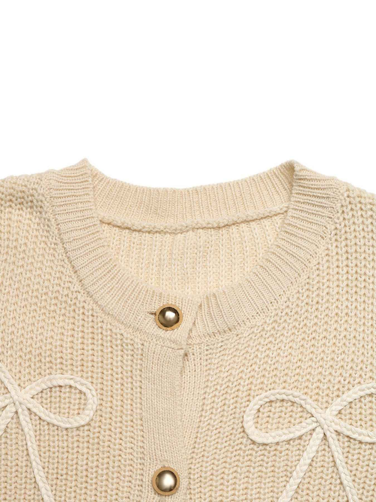 Close-up of a cream-colored knit cardigan sweater featuring a ribbed crew neck, gold-tone buttons, and decorative knitted bow details on the front, set against a plain white background.