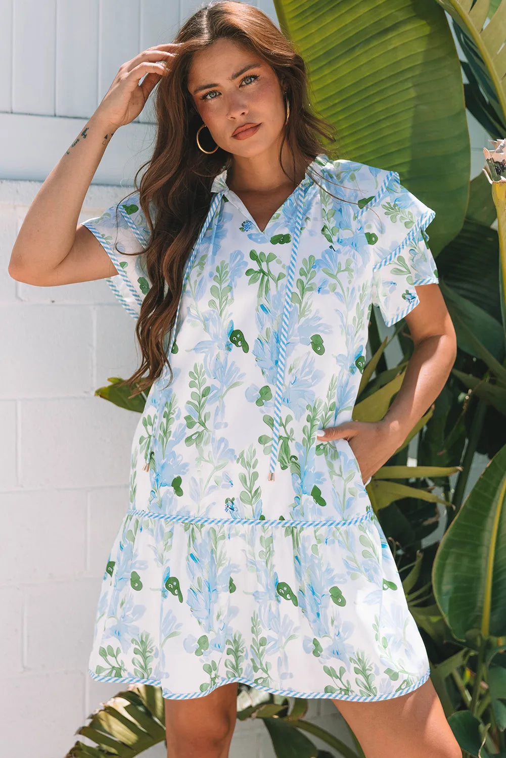 Woman wearing a floral dress standing in front of a white wall with green plants.