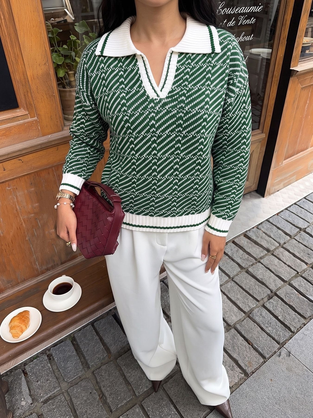 A woman wearing a green and white patterned knit polo sweater with white pants, holding a burgundy clutch bag, standing on a cobblestone street in front of a wooden storefront.
