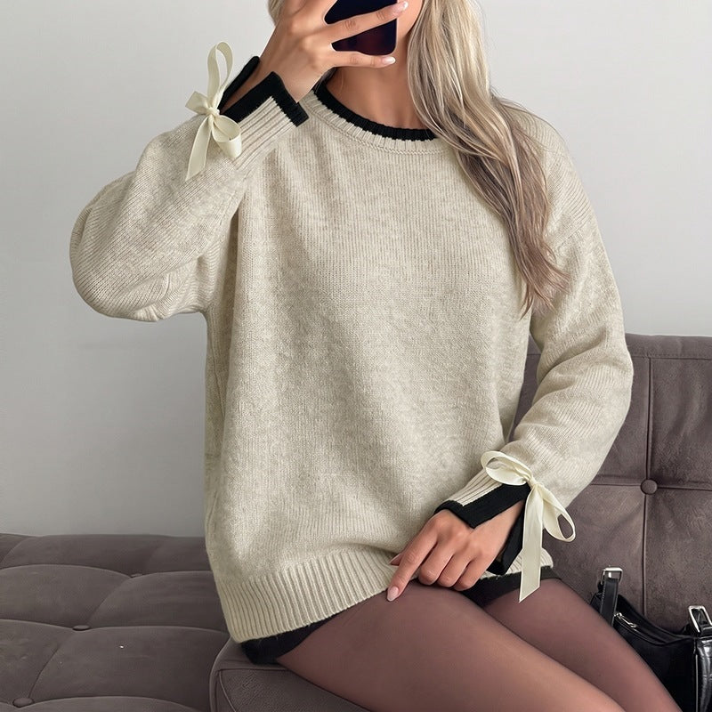 A woman wearing a cream-colored knit sweater with black trim at the collar and cuffs, featuring cream ribbon bows at the sleeve cuffs. She is sitting on a gray tufted sofa, taking a mirror selfie.