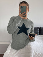 A woman takes a mirror selfie wearing a heather gray knit sweater with a large dark navy blue star on the chest, paired with white pants and holding a black mug.